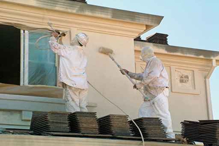 Deux travailleurs en combinaison qui peignent la façade d'une maison grâce à un rouleau à peinture et à un projecteur de peinture