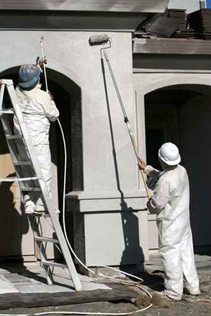 Deux travailleurs en combinaison qui peignent la façade d'une maison grâce à un rouleau à peinture et à un projecteur de peinture